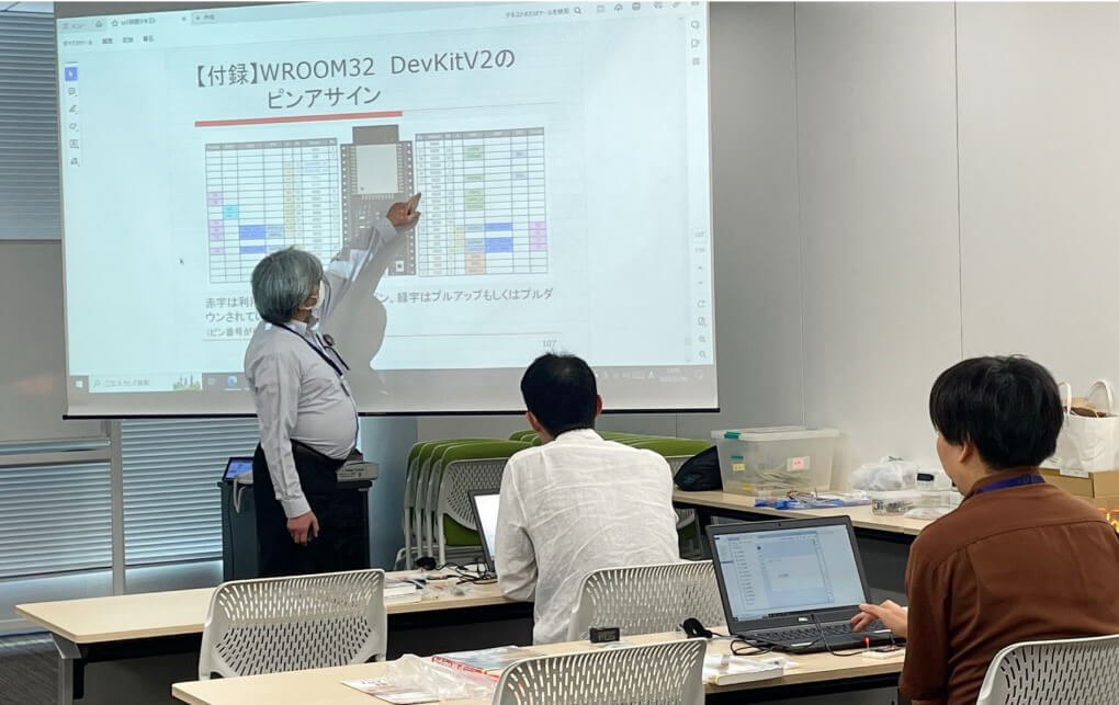 写真:研修風景。大きなスクリーンの前で講義を行う講師役の社員と、受講している社員の様子。