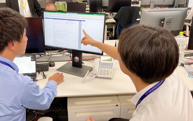 パソコンを指さし同僚と業務をしている増渕 仁史の後ろ姿の写真